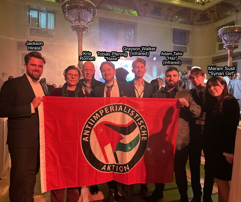 Several people standing behind a flag held by Hinkle and Haz that reads "Anti-Imperialische Aktion" and contains the combined Palestinian and Hammer and sickle symbols. Most of the people are identified in the surrounding text.